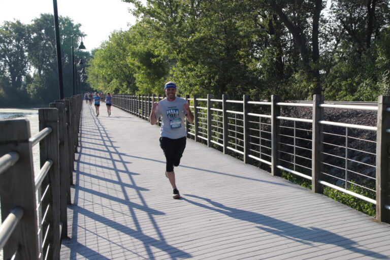 Man running on bridge in ThedaCare Half Marathon with trees and blue sky.